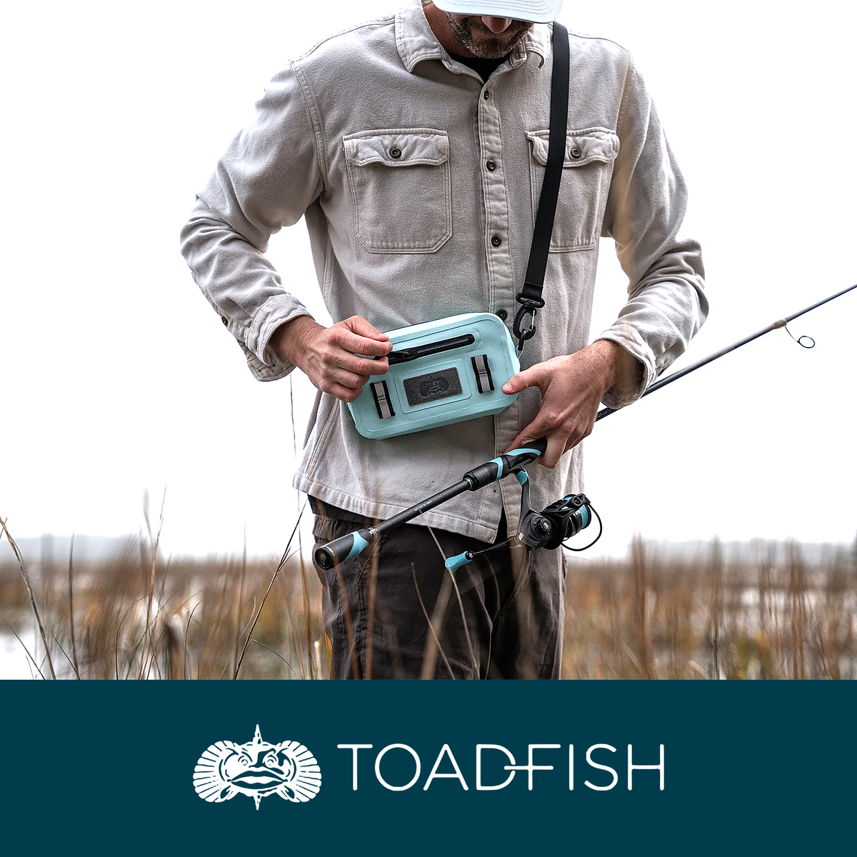 A man standing in marsh grass holding a Toadfish Sealed Sling tackle bag and a fishing rod, above the Toadfish wordmark on a taupe band.