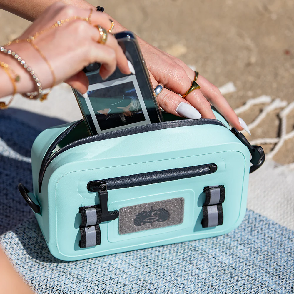 Phone being placed into the Seafoam Toadfish Sealed Sling on a beach blanket